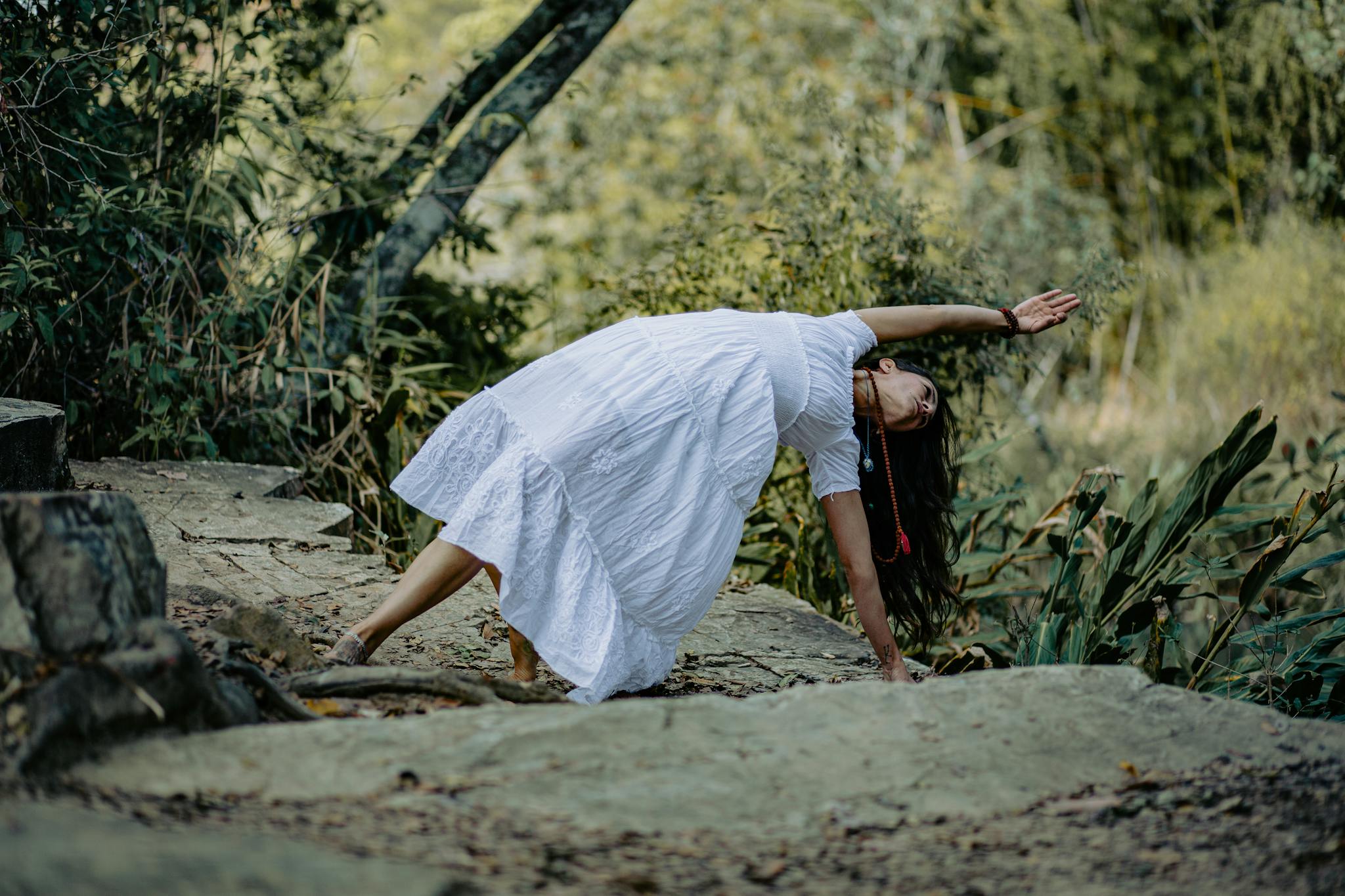 Woman in white dress practicing yoga on stone path in a lush forest setting.